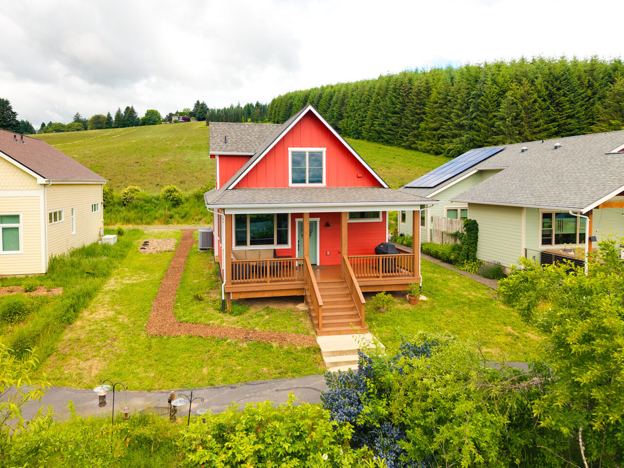 The red farmhouse-style home with a wide covered front porch, steps down to a path, with a green hill and fir trees rising behind it.