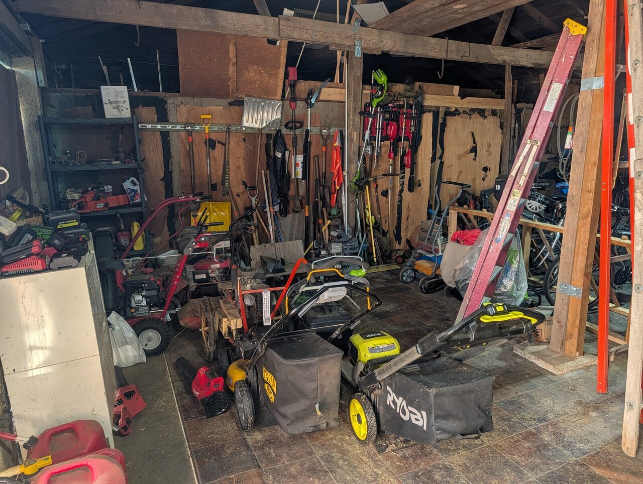 Mowers, ladders and hand tools racked on the wall of a timber-framed shed.